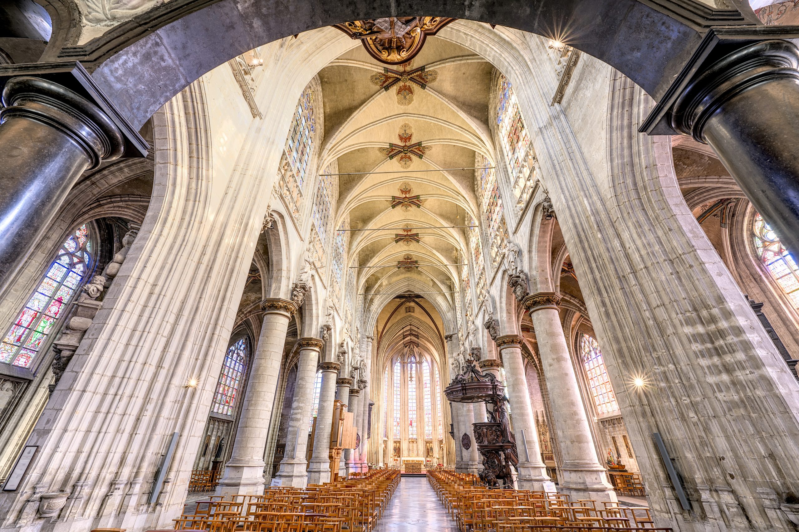Vue intérieure de la nef de l'église éclairée avec des colonnes, un plafond élevé et des fenêtres colorées.