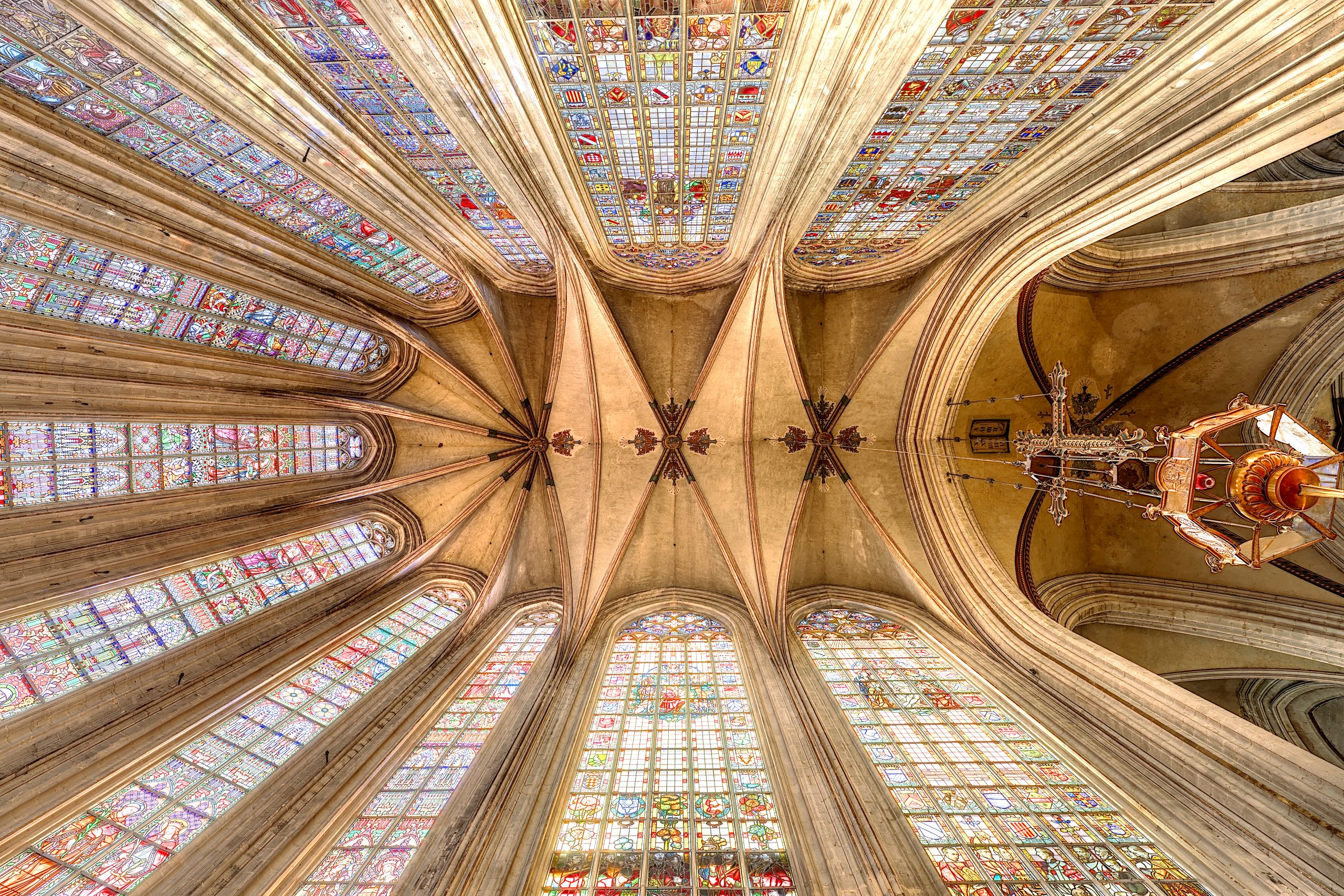 Vue du plafond voûté de l'église Notre-Dame au Sablon, avec des vitraux colorés et un grand lustre suspendu.